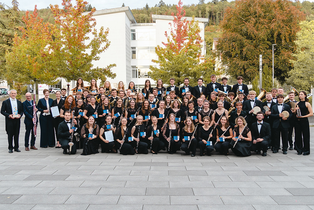 Große Gruppe von Orchestermitgliedern in schwarzer Konzertkleidung auf einem Vorplatz vor einem modernen Gebäude. Die Musikerinnen und Musiker halten verschiedene Blas- und Schlaginstrumente sowie Auszeichnungen in den Händen. Im Hintergrund sind herbstlich gefärbte Bäume zu sehen.